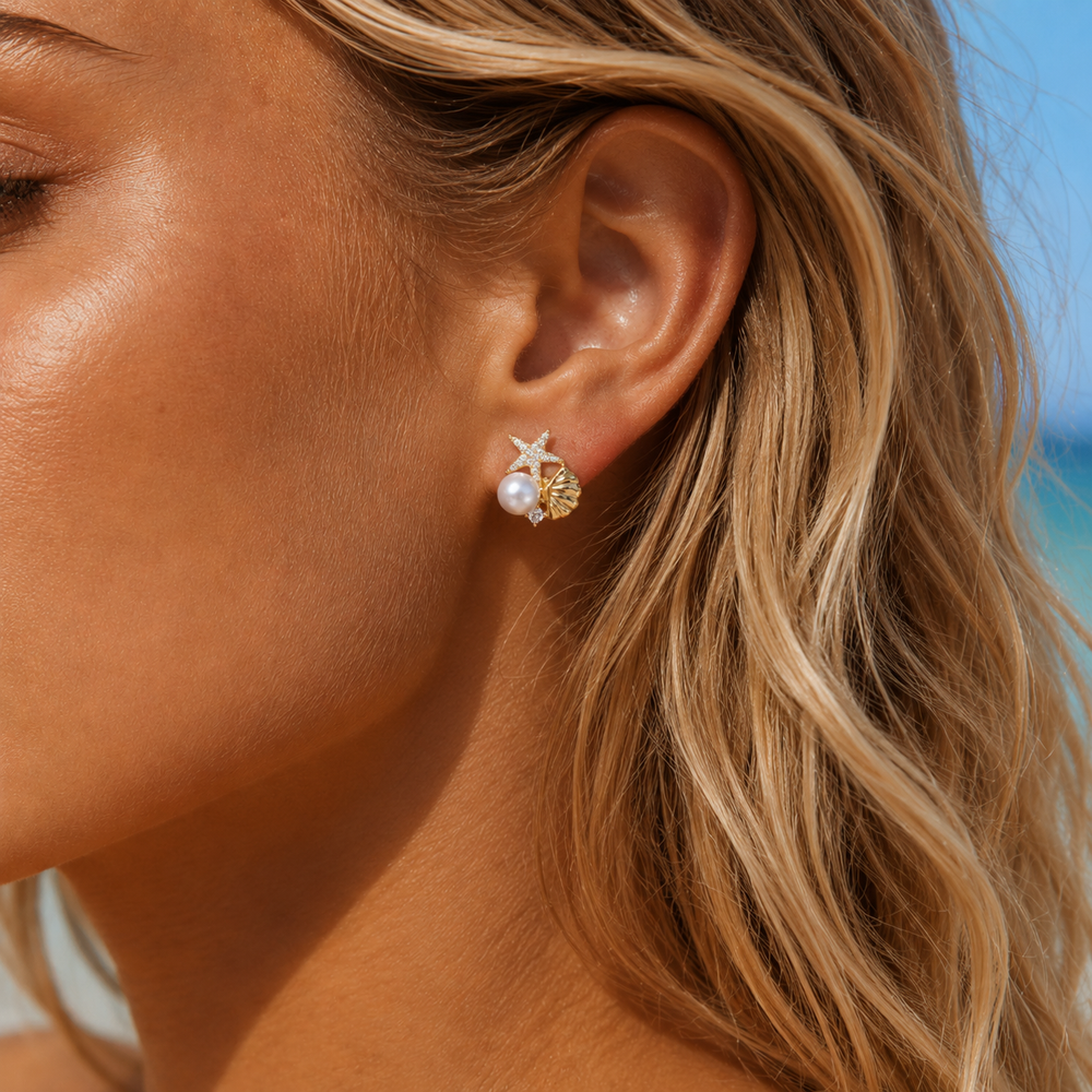 Close-up of a woman's ear wearing gold and pearl earrings with a blurred beach background.